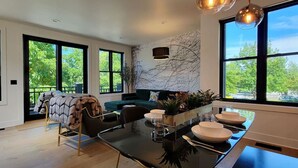 Eine moderne Küche mit einem Glastisch, einer Couch und Blick auf Bäume durch große Fenster.