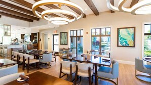 Un vaste espace de salle à manger avec de nombreuses tables et chaises, un coin cuisine avec des tabourets de bar, et de grandes fenêtres qui laissent entrer la lumière naturelle et offrent une vue sur l’extérieur.