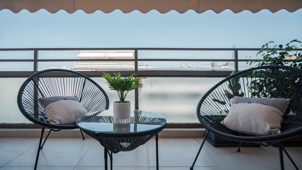 A balcony with two wicker chairs and a small table, a potted plant, and a view of a bridge and buildings across the water. - Modern Apartment with Acropolis view (Athens)