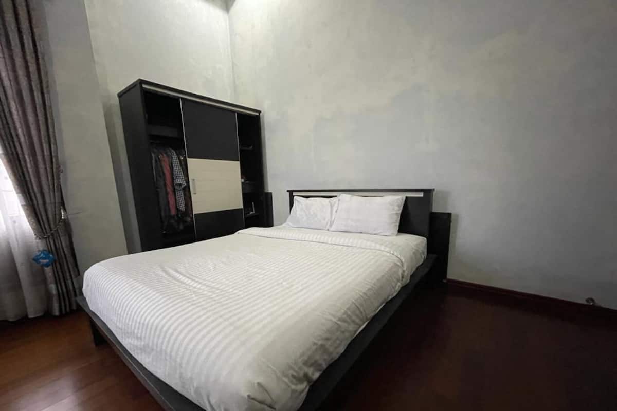 A neatly made bed with a white bedspread and pillows, a dark headboard, and a wardrobe with a mirror and hanging clothes.
