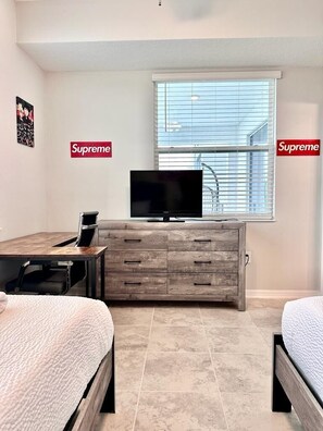 Een slaapkamer met een bed, een bureau, een televisie op een houten dressoir en een raam met jaloezieën.