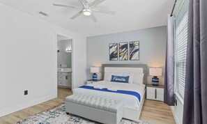 Ein Schlafzimmer mit Bett, Nachttischen, Deckenventilator, Kunstwerken und einem durchgehenden Türrahmen sichtbarem Badezimmer.