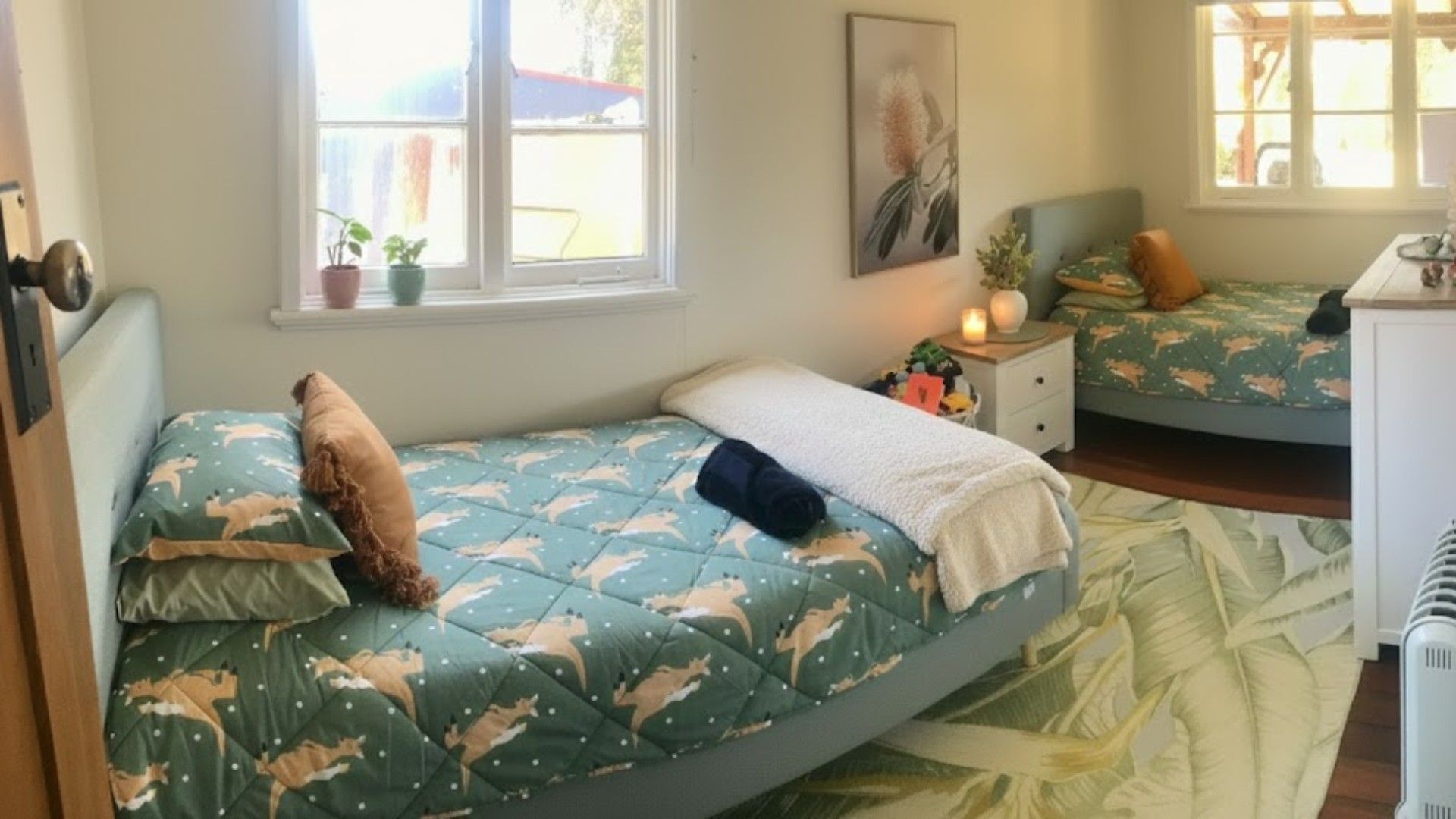 A bedroom with two beds, each with a patterned bedspread and pillows. There are potted plants on the windowsill and a painting on the wall.