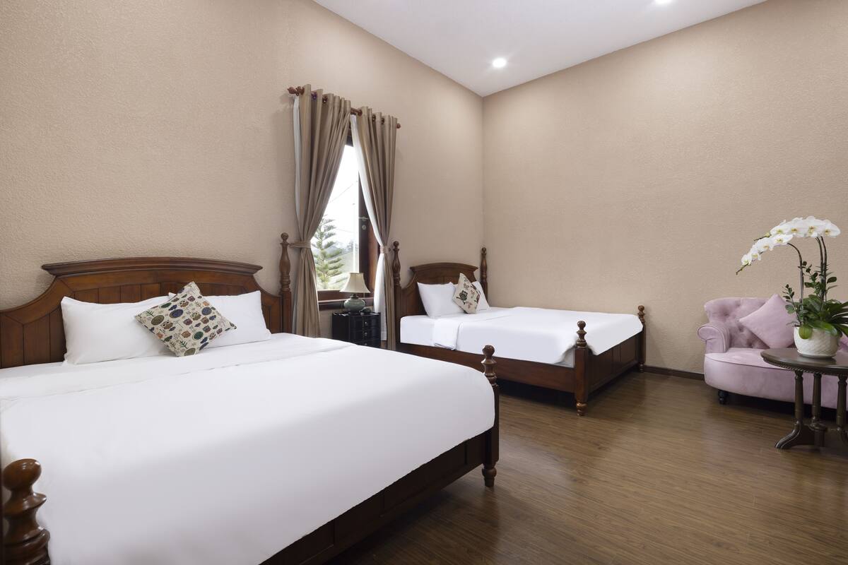 A hotel room with two wooden beds, a small pink armchair, and a table with a potted plant.