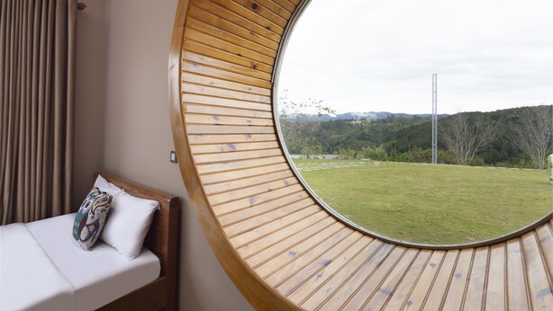 Ein Schlafzimmer mit einem runden Holster, einem Bett mit weißer Matratze und einem Zierkissen, und Blick auf eine grüne Landschaft mit Bäumen und Bergen.