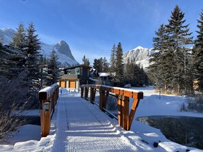 A wooden bridge leading to a modern building with large windows, surrounded by snow-covered trees and mountains.