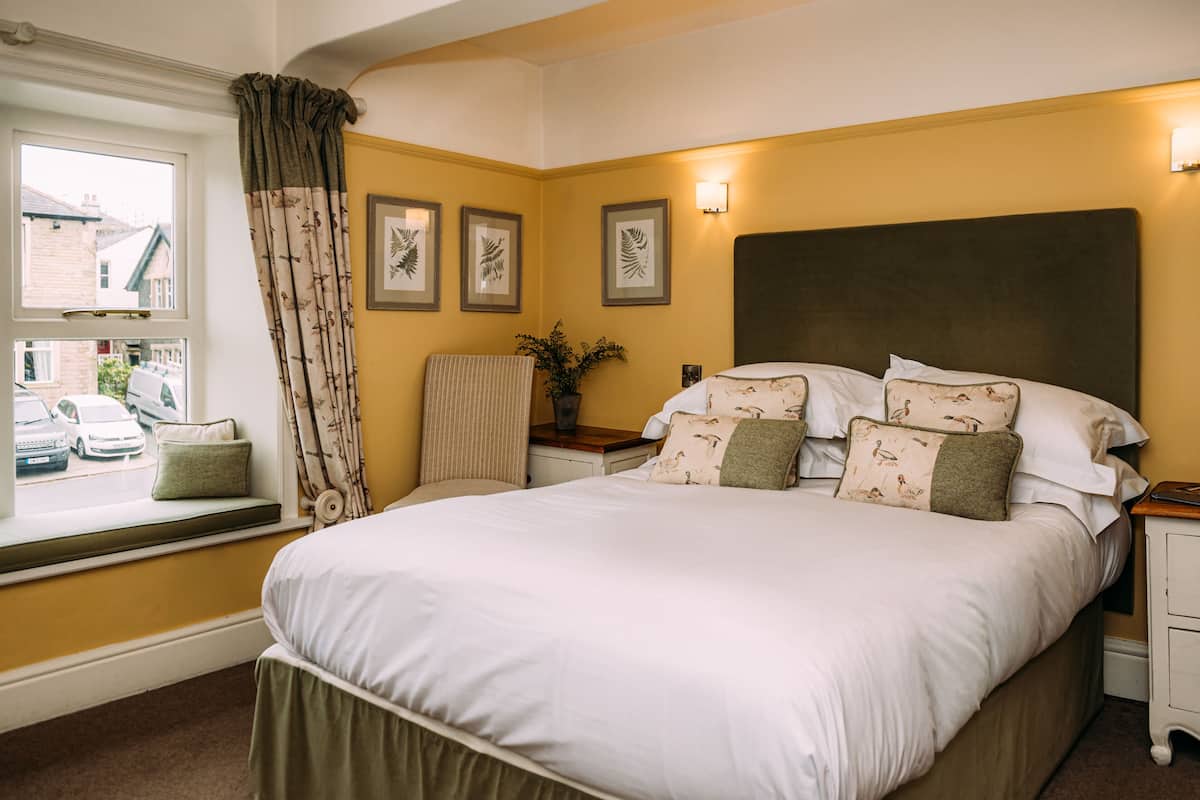 A neatly made bed with white linens and multiple pillows, a bedside table with a lamp, and a window with a view of the street.