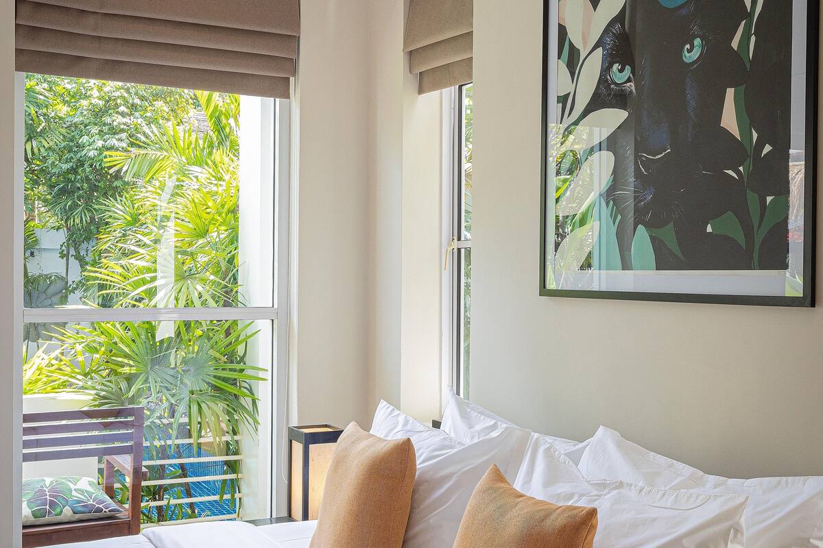 Hotelkamer met een bed, witte kussens, een groot raam met uitzicht op groen en een lijst met een kunstwerk aan de muur.