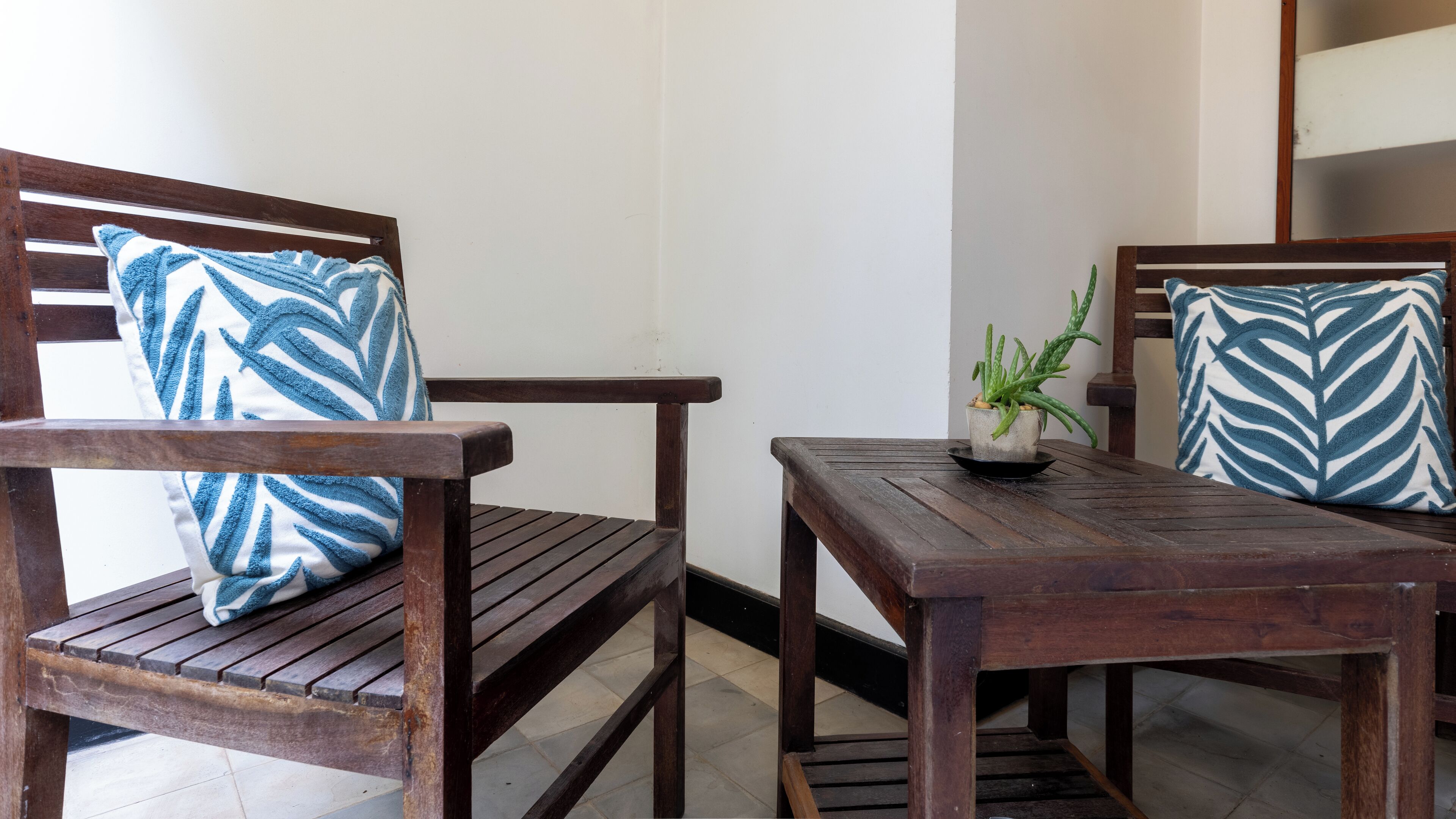 Un banco de madera con un cojín de diseño azul y blanco, una mesita con una planta en maceta, y otro banco con un cojín similar.