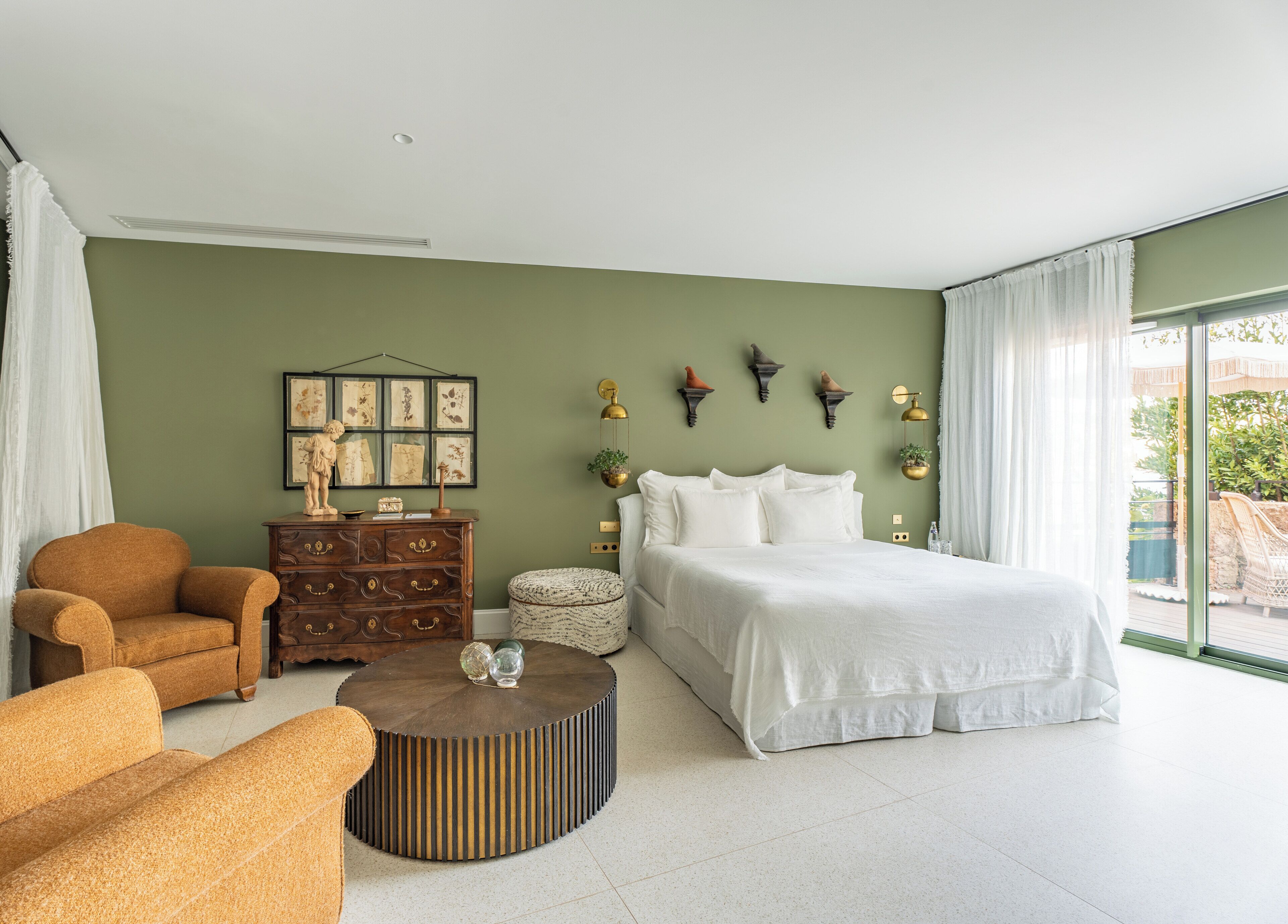 Ein modernes Schlafzimmer mit einem großen Bett, einer hölzernen Kommode, einem runden Couchtisch und Blick ins Freie durch ein großes Fenster.