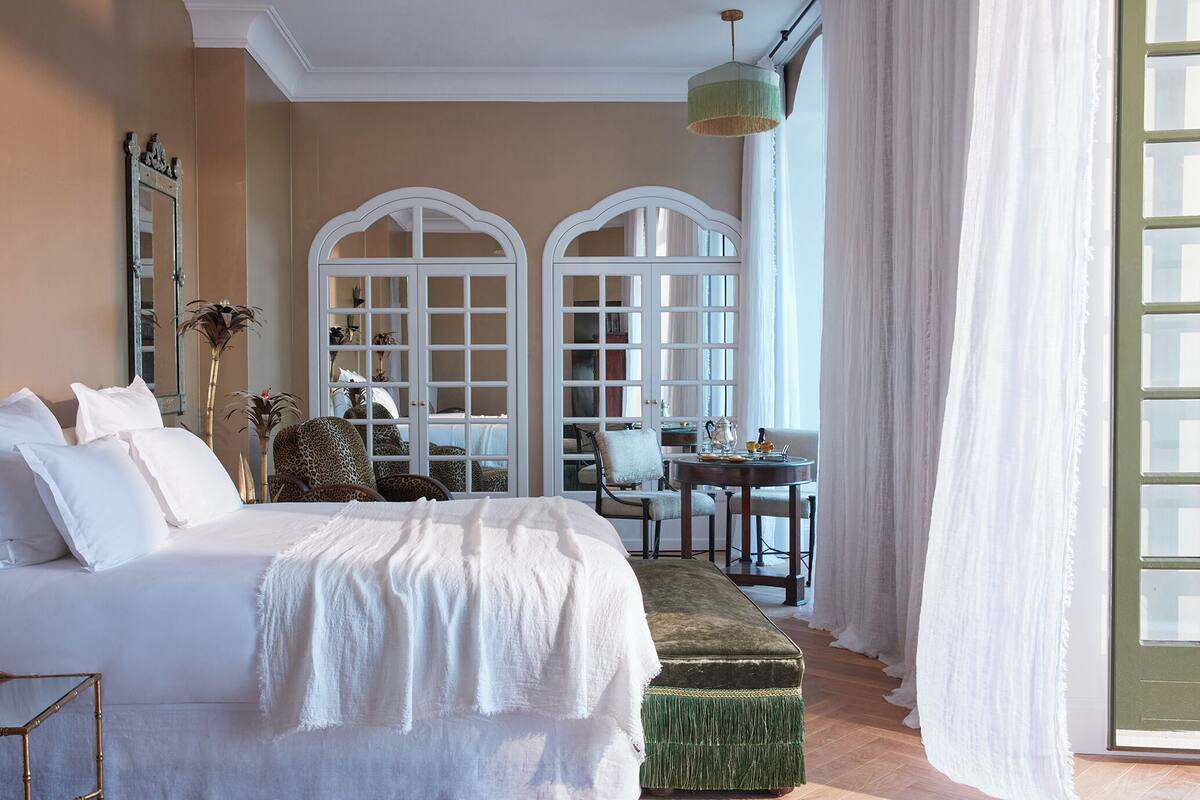 Ein Schlafzimmer mit einem großen Bett, einem grünen Pouf, einem Schreibtisch, Stühlen und einem Spiegel.