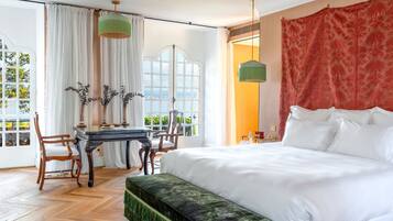Habitación de hotel con una cama grande, una mesa de comedor con sillas, un banco y vistas a zonas verdes por la ventana.