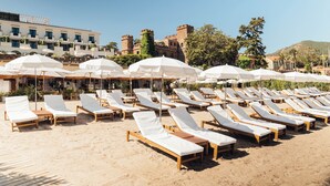 Una playa con filas de tumbonas y sombrillas, un edificio que recuerda a un castillo al fondo, y un cielo despejado.