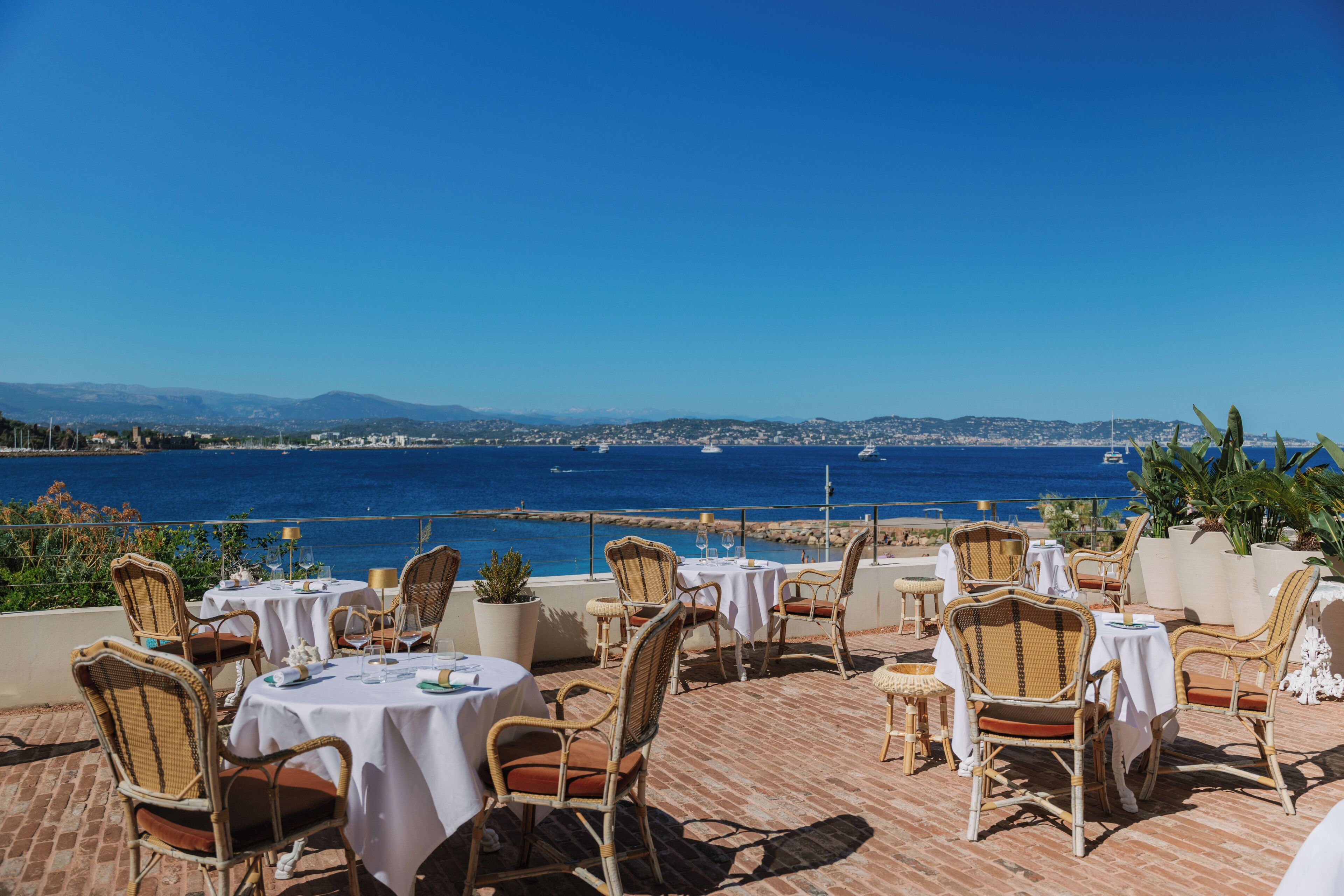 A terrace with wicker chairs and tables, overlooking a coastal view with boats and distant mountains.