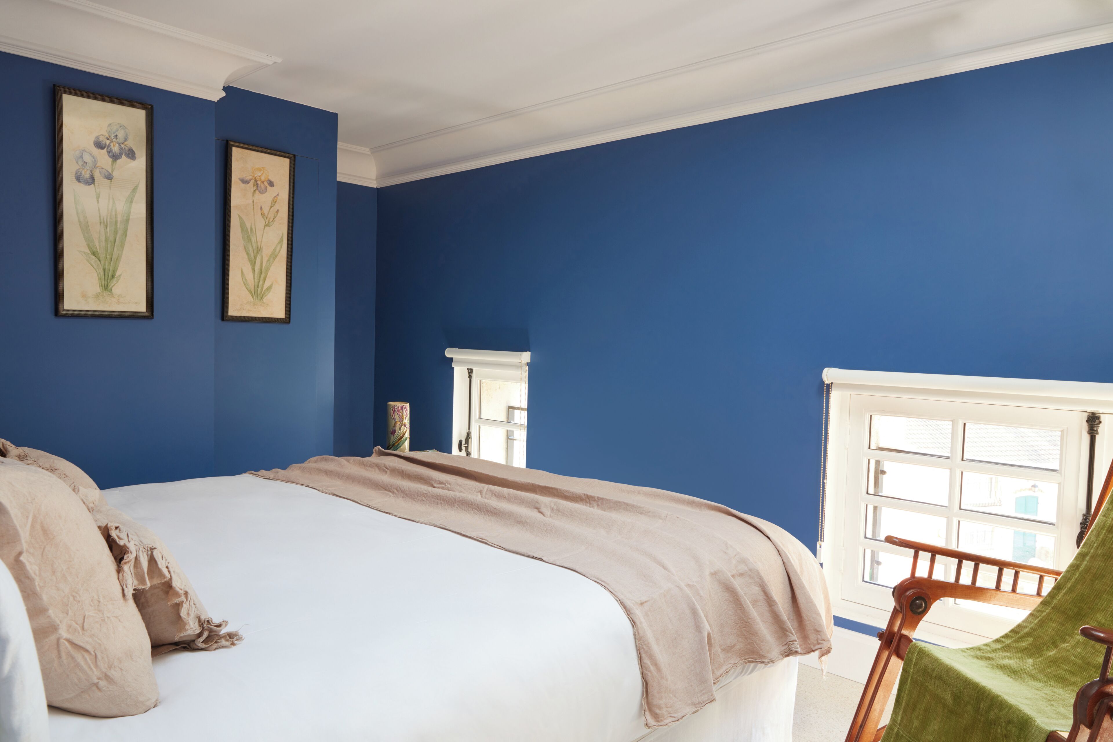Un dormitorio con una cama grande, paredes azules, una ventana y dos cuadros florales enmarcados.