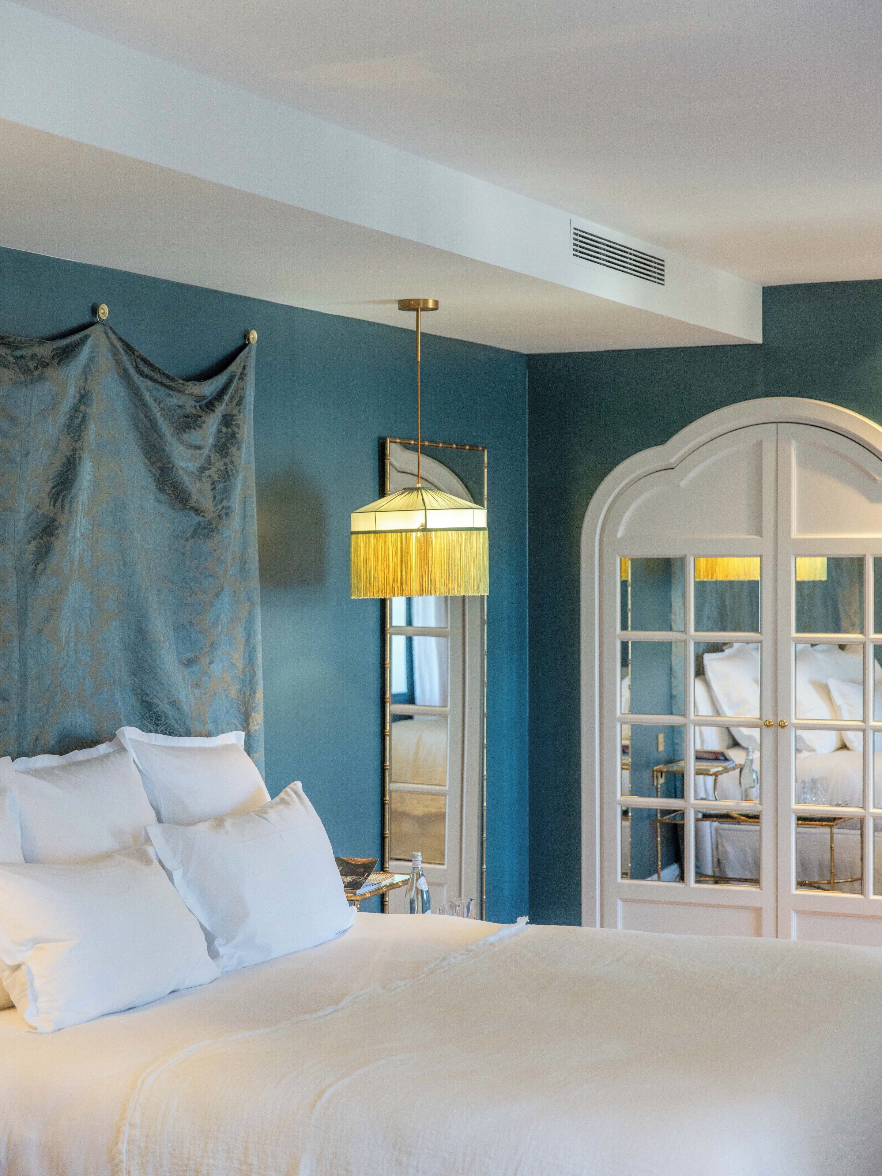 A bedroom with a bed, white curtains, a hanging lamp, and a mirror with a white frame.