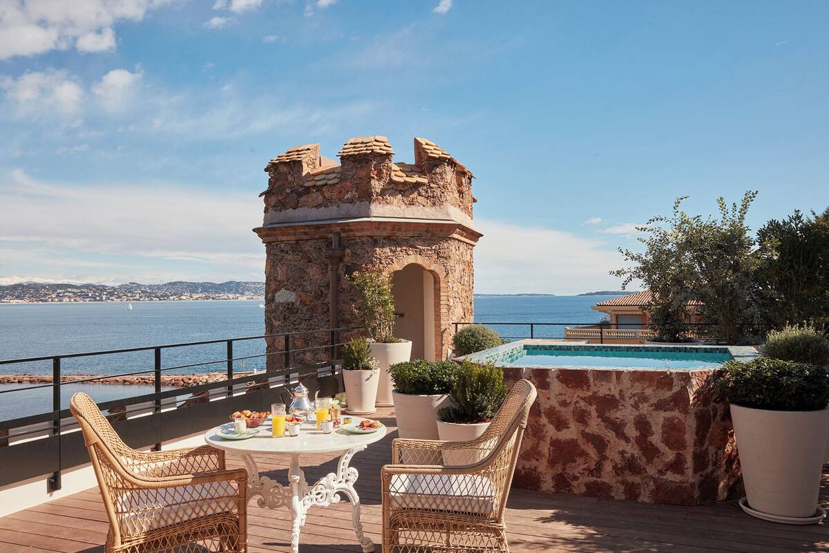 Eine Terrasse mit Korbstühlen, einem Tisch mit Frühstücksgebäck, einem Steinturm und einem Pool.