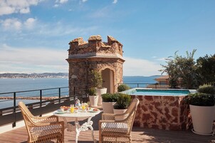 A terrace with wicker chairs, a table with breakfast items, a stone tower, and a pool.