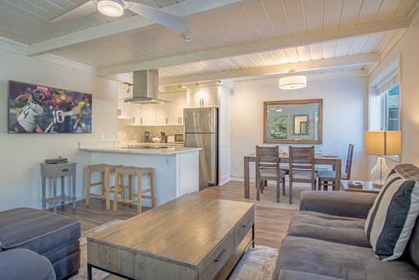 A modern kitchen with a central island, wooden stools, a dining table with chairs, a sofa, and a large painting on the wall.