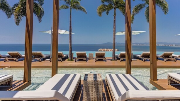 Un espace aménagé au bord de la piscine, avec des chaises longues et des parasols, offrant une vue sur une mer d’un bleu limpide et sur des montagnes lointaines.