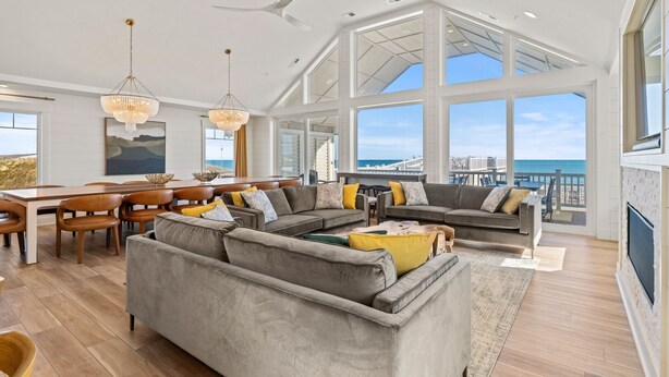 A spacious living room with a large gray sectional sofa, a dining area with wooden chairs, and a view of the ocean through large windows. - Island Enchantment - Oceanfront Pet Friendly w/ Cabana Service (Kill Devil Hills)