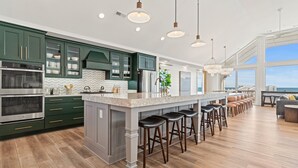 A modern kitchen with dark green cabinets, stainless steel appliances, and a large island with bar stools. - Island Enchantment - Oceanfront Pet Friendly w/ Cabana Service (Kill Devil Hills)