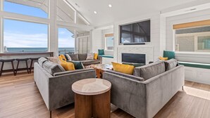 A spacious living room with a large window overlooking the sea, a fireplace, and a cozy seating area. - Island Enchantment - Oceanfront Pet Friendly w/ Cabana Service (Kill Devil Hills)