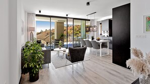 Un salon moderne doté d’une grande porte-fenêtre coulissante donnant sur un balcon, d’un coin repas avec une table et des chaises blanches, et d’un espace cuisine avec un meuble noir et des luminaires suspendus.