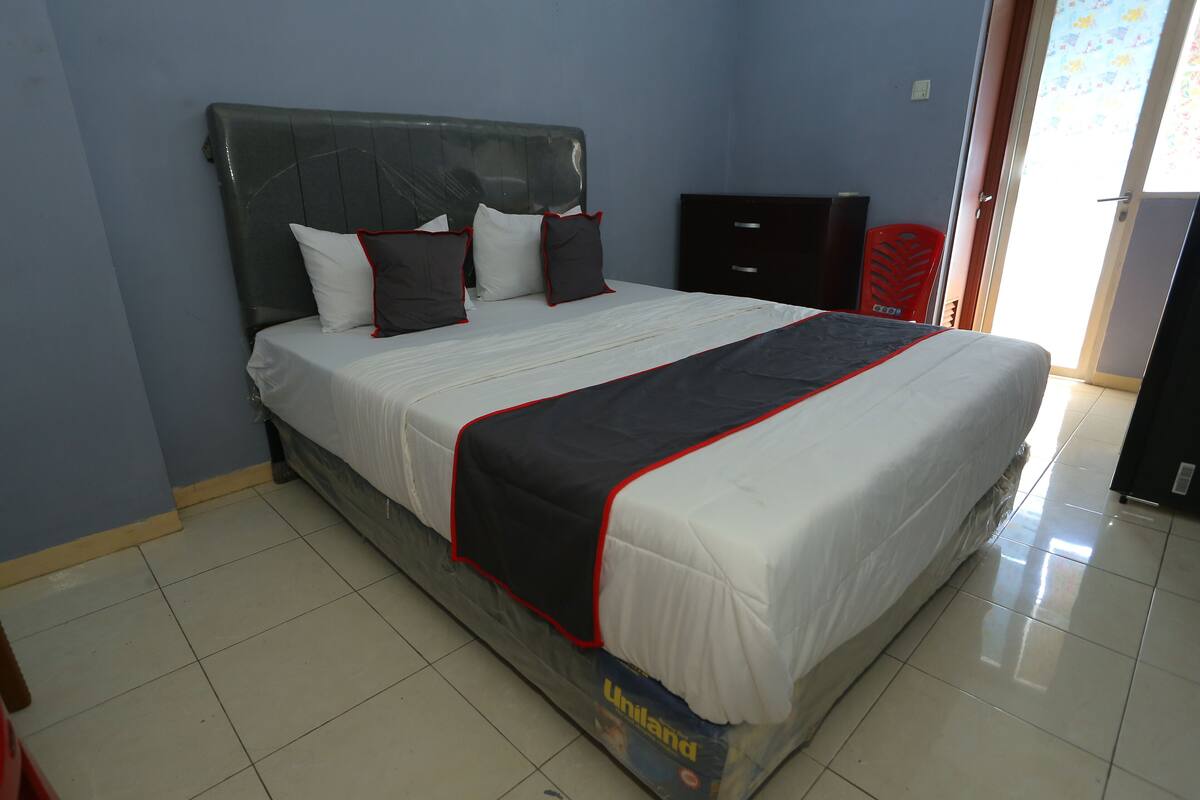 A neatly made bed with a black and white bedspread and red trim, a dark headboard, and a red chair in a room with light blue walls and a tiled floor.