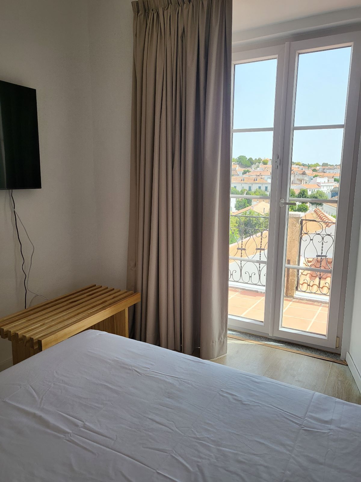 Un dormitorio con cama, una banca de madera, televisor montado en la pared y puerta corrediza de vidrio que da a un balcón con vistas a edificios.