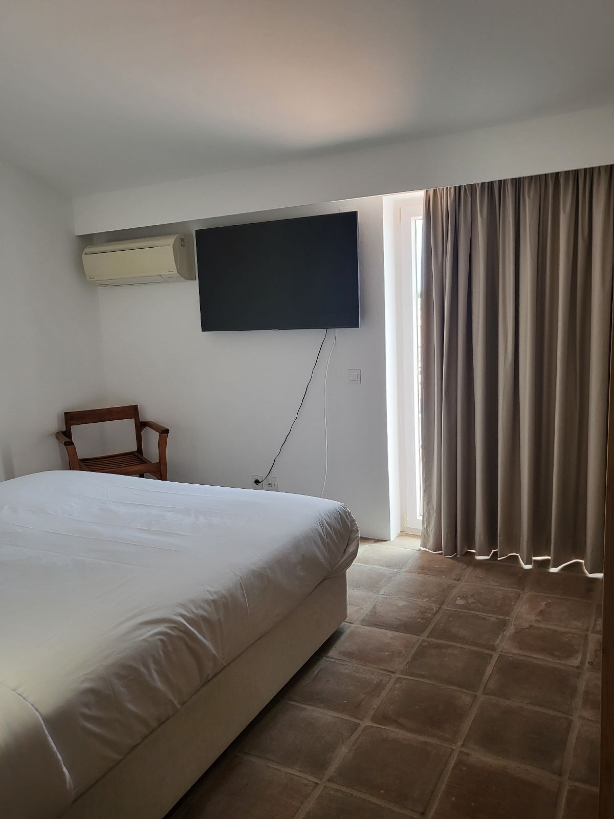 A bedroom with a large bed, a flat-screen TV mounted on the wall, a wooden chair, and a window with curtains.