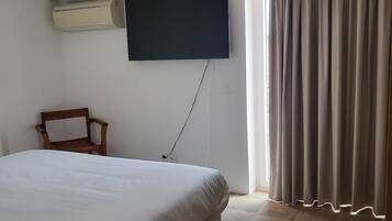 A bedroom with a large bed, a flat-screen TV mounted on the wall, a wooden chair, and a window with curtains.