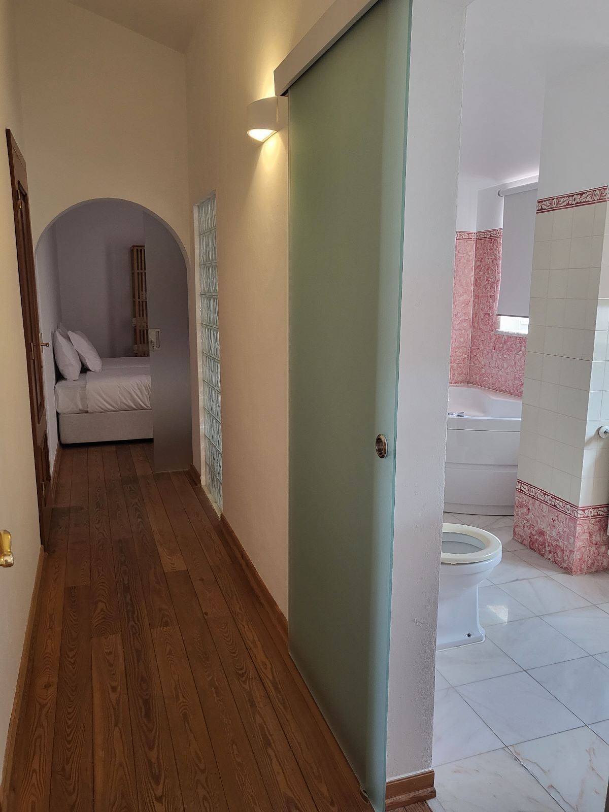 Een gang die leidt naar een slaapkamer met een bed, een deur en een badkamer met bad en toilet.