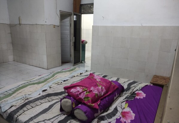 A small, sparsely furnished room with a bed, a small stool, and a door leading to another room.