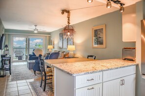 A kitchen with a large island, white cabinetry, and a chandelier. There is a dining area with chairs and a table, and a balcony with a view.
