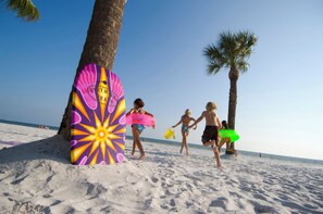 A beach scene with people walking and holding surfboards, palm trees, and a colorful beach umbrella.