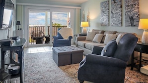 Un salon comprenant un canapé, des fauteuils, une table basse et un téléviseur. Il y a un balcon avec une balustrade et une vue sur le ciel.