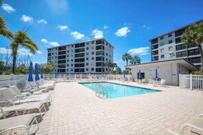 A swimming pool with lounge chairs and umbrellas, surrounded by a white fence, with multi-story buildings in the background.