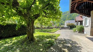 A stone pathway leading to a building with a red-tiled roof, surrounded by lush greenery and a tree.
