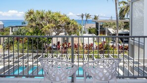 Ein Balkon mit zwei weißen Stühlen, einem Swimmingpool und Blick auf das Meer und Palmen.