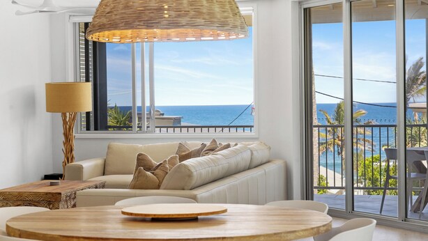 Ein modernes Wohnzimmer mit einer Couch, einem runden Holztisch und einem großen Fenster mit Blick auf das Meer.
