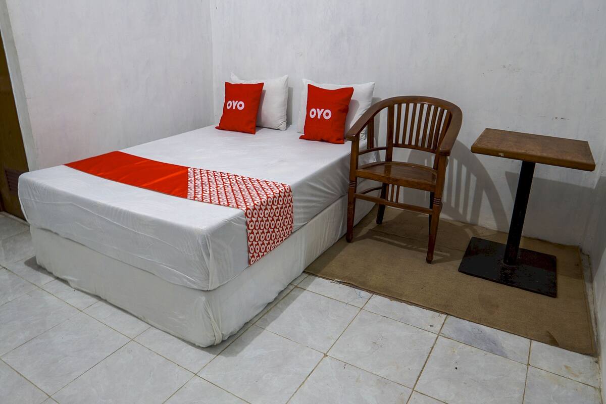 Una cama individual con colchón blanco y detalles rojos, una silla de madera y una mesita con base negra.