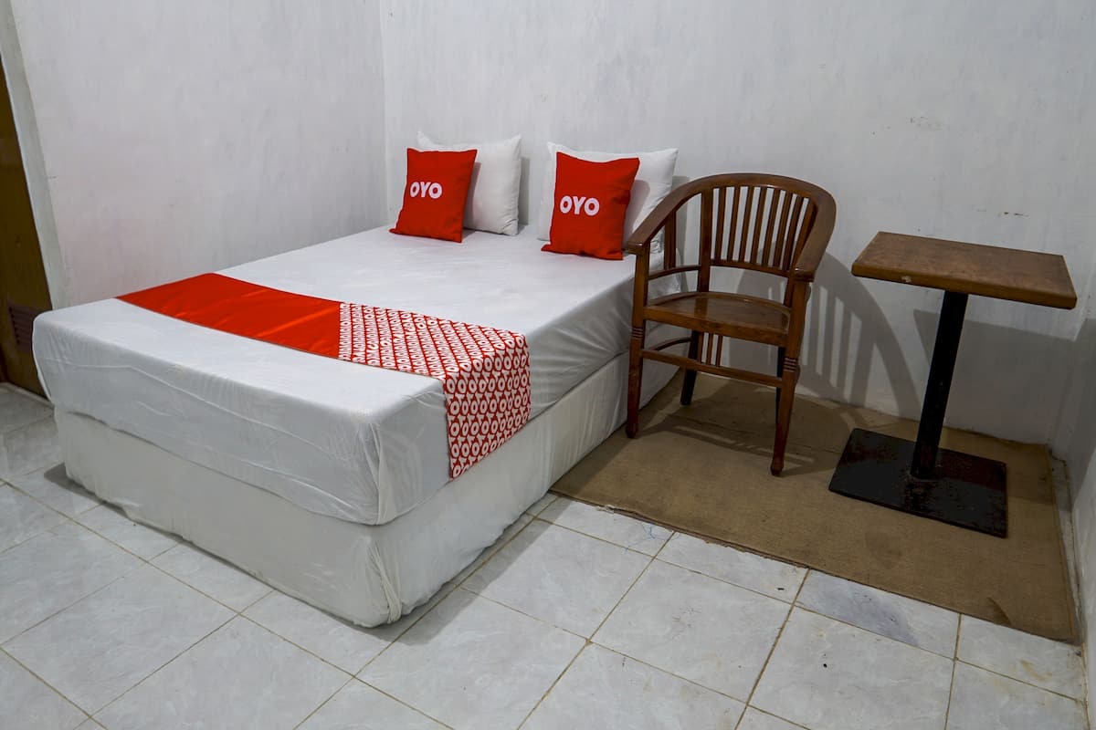Una cama individual con colchón blanco y detalles rojos, una silla de madera y una mesita con base negra.