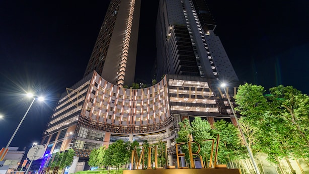 Um edifício moderno com fachada curva, iluminado por postes de luz, cercado por vegetação e uma placa com a inscrição 