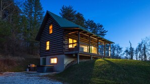 A wooden cabin with a covered porch, a fireplace, and a view of the surrounding forest.