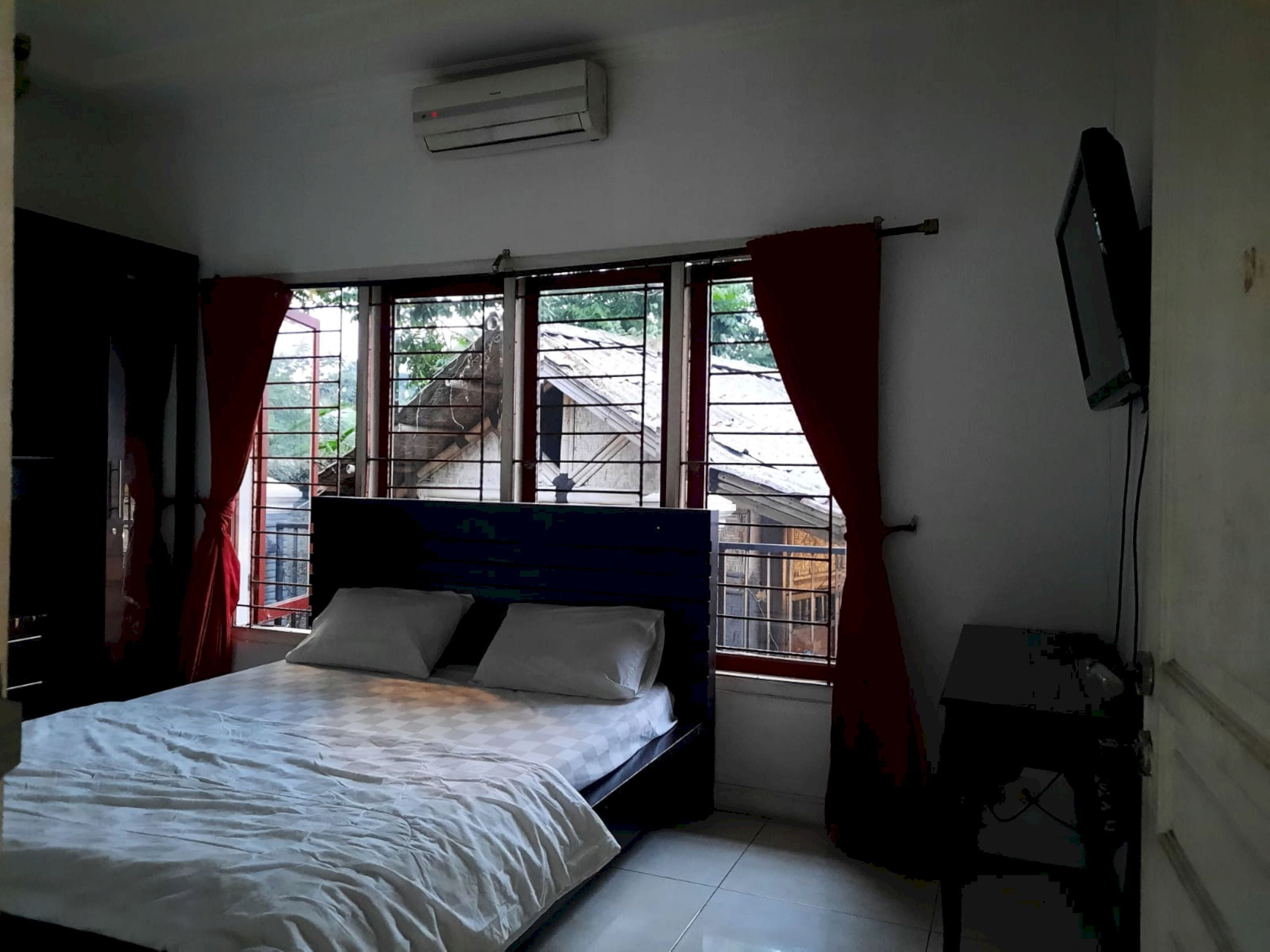 Ein Schlafzimmer mit Bett, einem Fenster mit Vorhängen, einem Fernseher an der Wand und einer Klimaanlage.