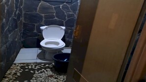 A small bathroom with a stone wall, a white toilet, and a blue bucket. - OYO 93159 Resort Cinta Kasih (Megamendung)
