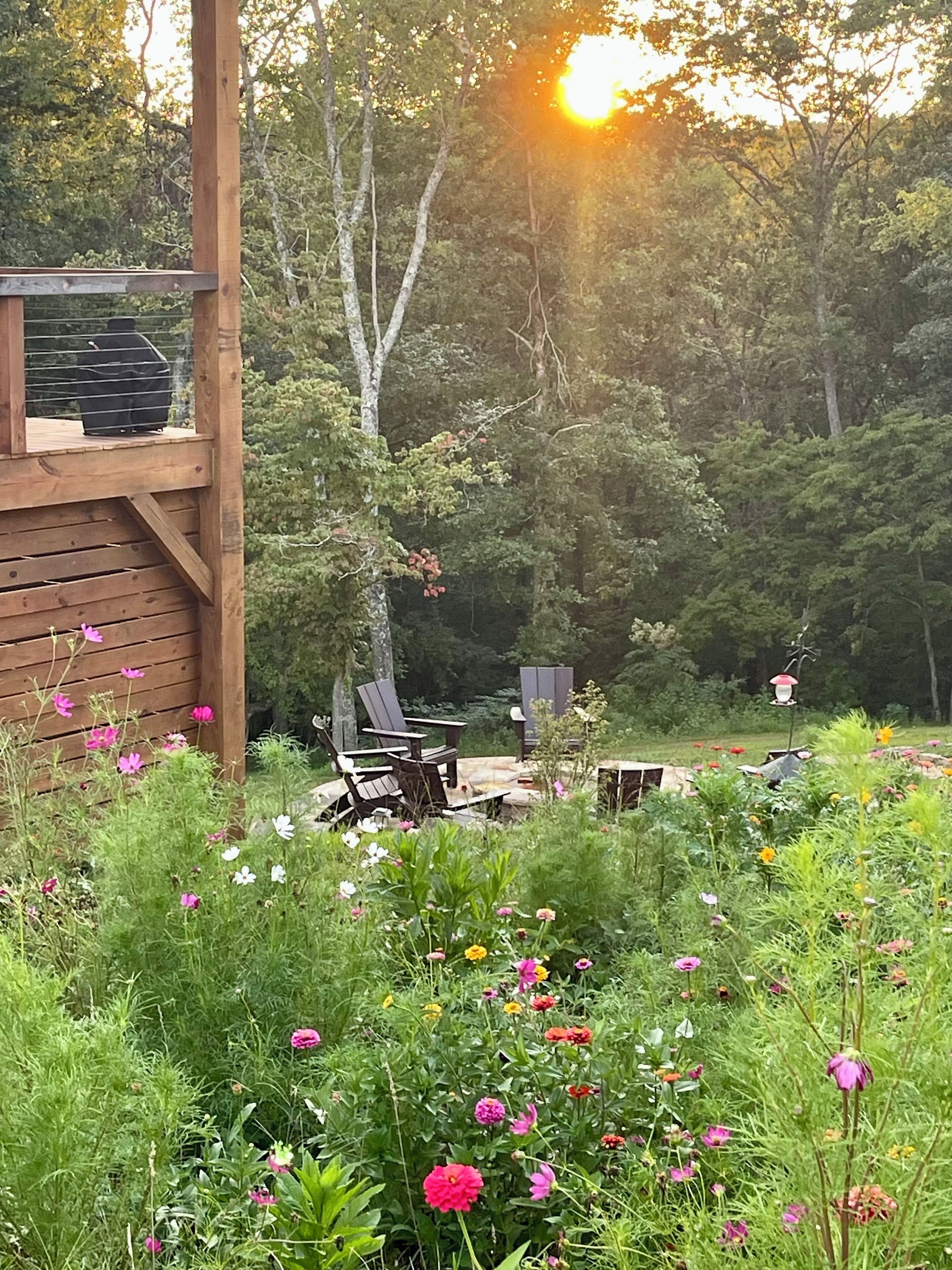 Una terraza de madera con mobiliario exterior, rodeada por un jardín con diversas flores y árboles.