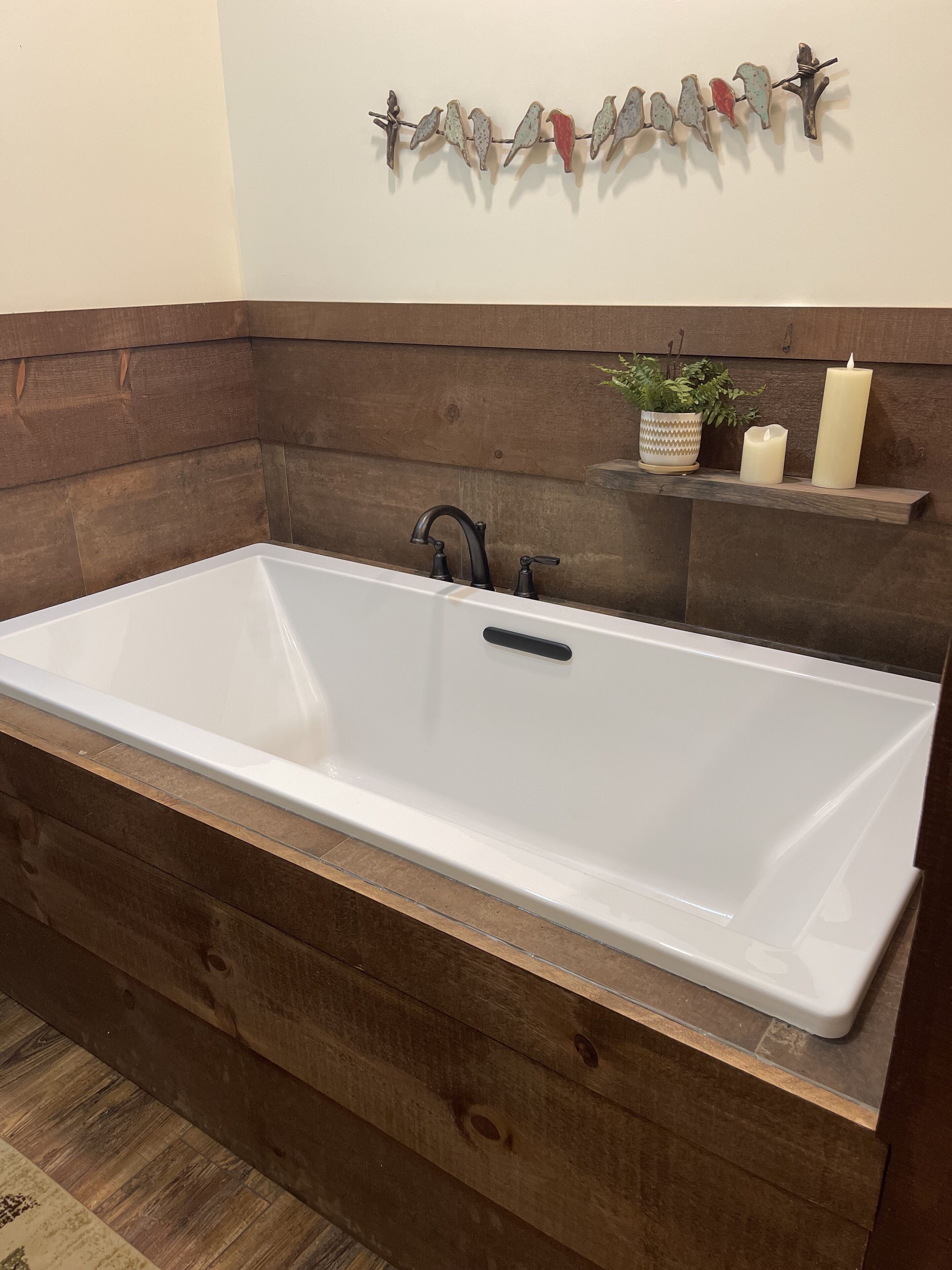 Una bañera rectangular con base de madera, un estante en la pared con una planta y velas, y una decoración de pájaro en la pared.