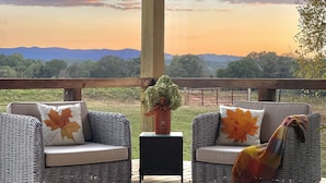 A covered outdoor seating area with wicker furniture and cushions, a small table, and a view of distant mountains and trees. - The Dogwood Inn (Blue Ridge)